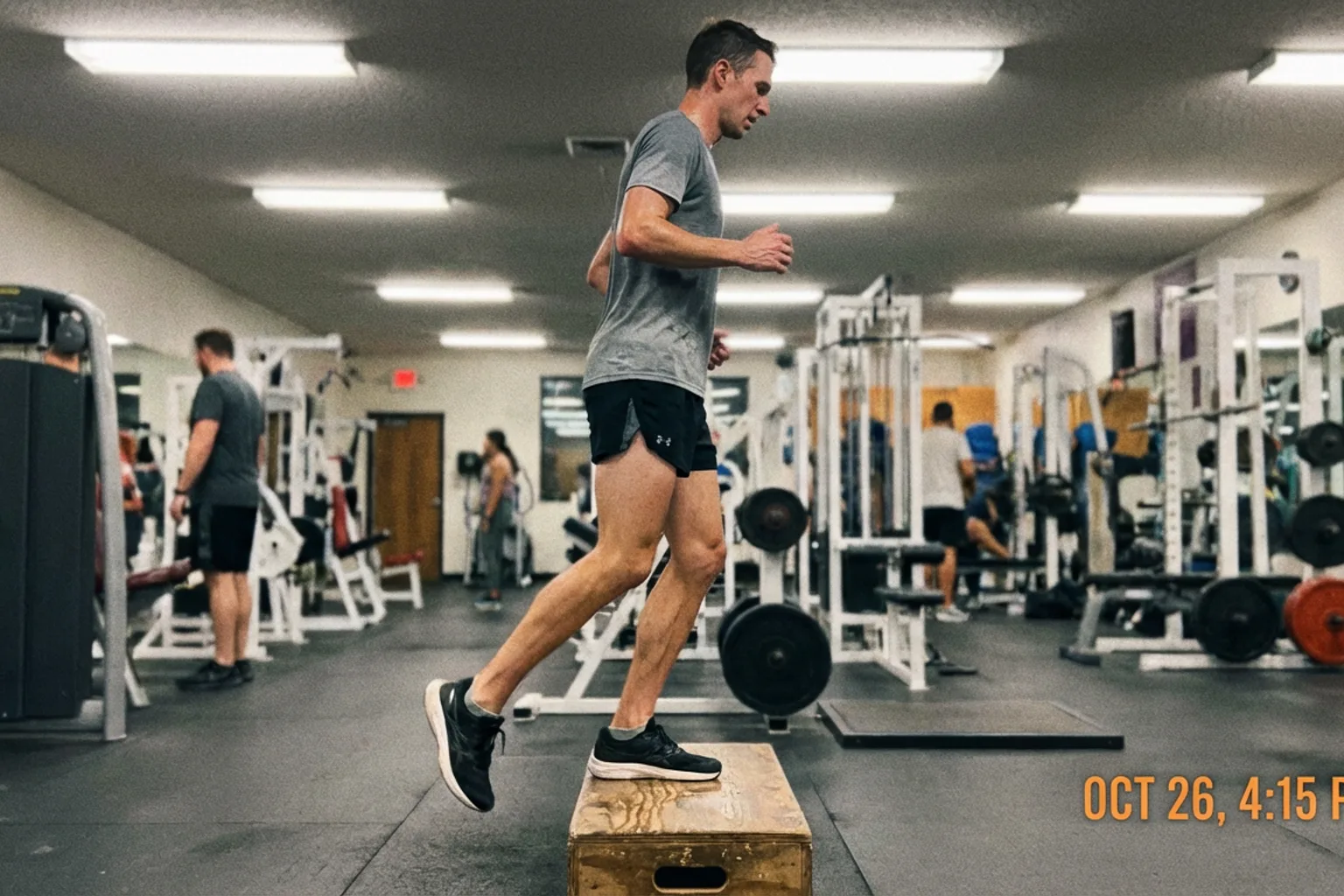 Candid iPhone photo of a lean male runner doing single-leg calf raises on a wooden gym step, natural overhead gym lighti
